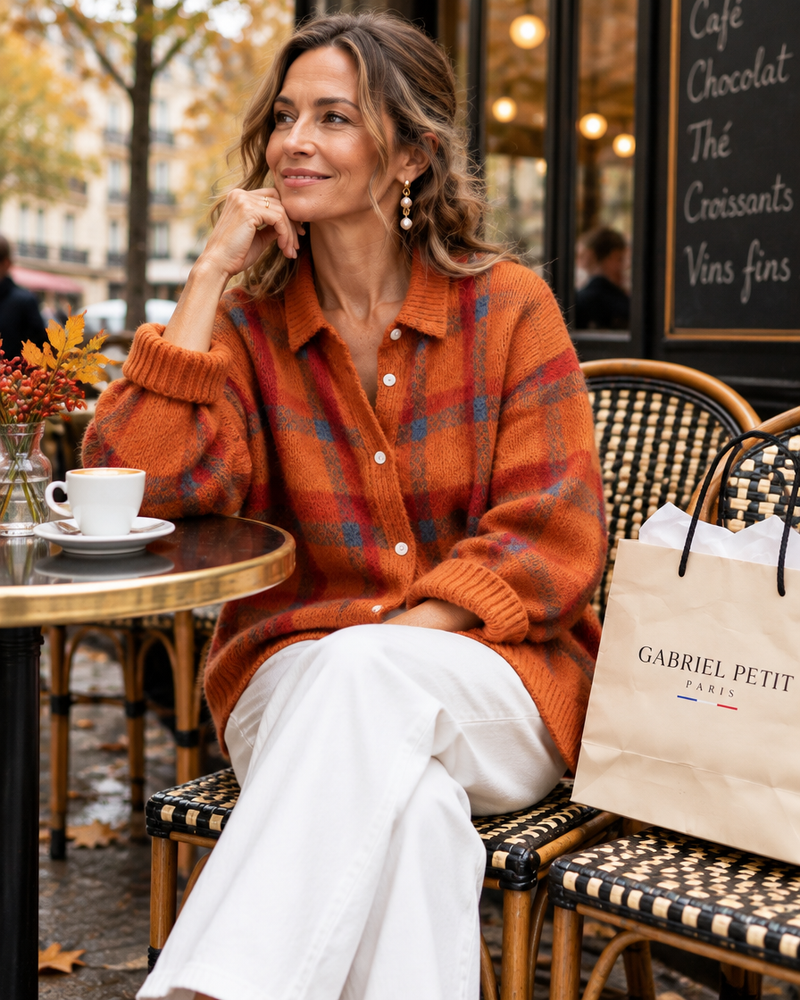 Femme d'une cinquantaine d'années en cardigan orange à carreaux et pantalon blanc, assise en terrasse à Paris, avec un sac GabrielPetit